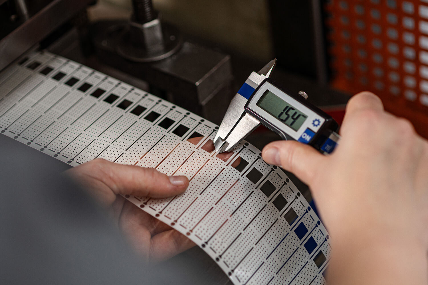 Nahaufnahme der Hände eines Mitarbeiters, der mit einem digitalen Messschieber ein präzise gestanztes Folienbauteil vermisst.