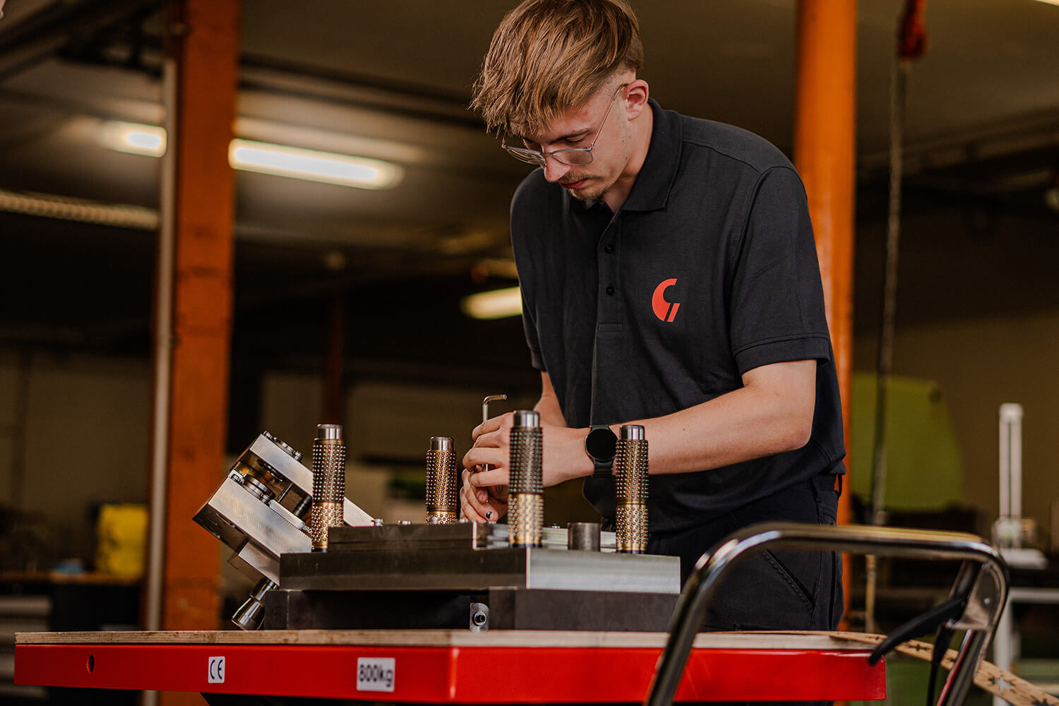 Ein junger Mitarbeiter im Werkzeugbau montiert konzentriert ein mehrteiliges Stanzwerkzeug auf einer roten Werkbank.