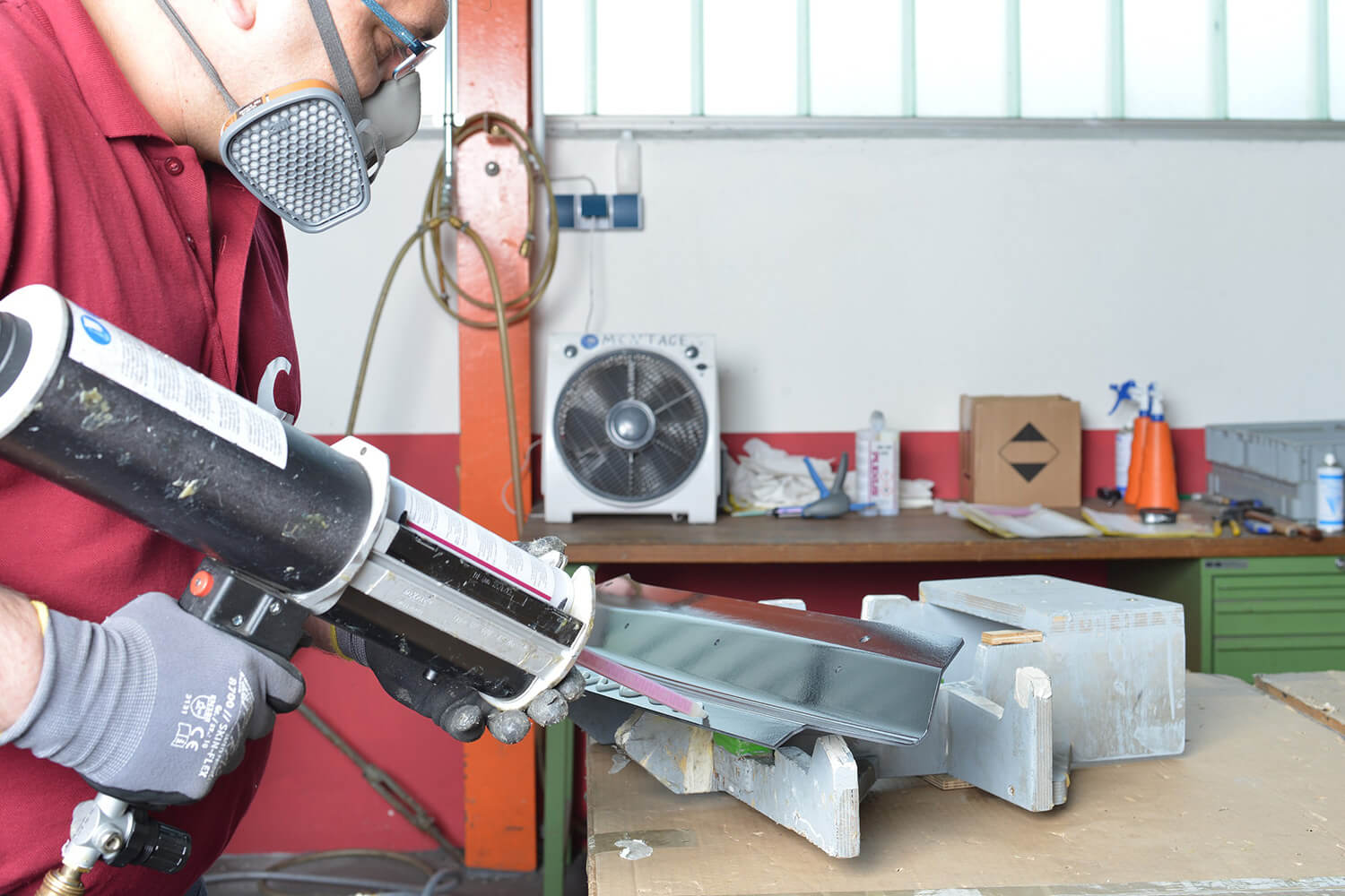 Ein Mitarbeiter mit Atemschutzmaske trägt mit einer professionellen Kartuschenpresse Klebstoff auf ein Bauteil auf.